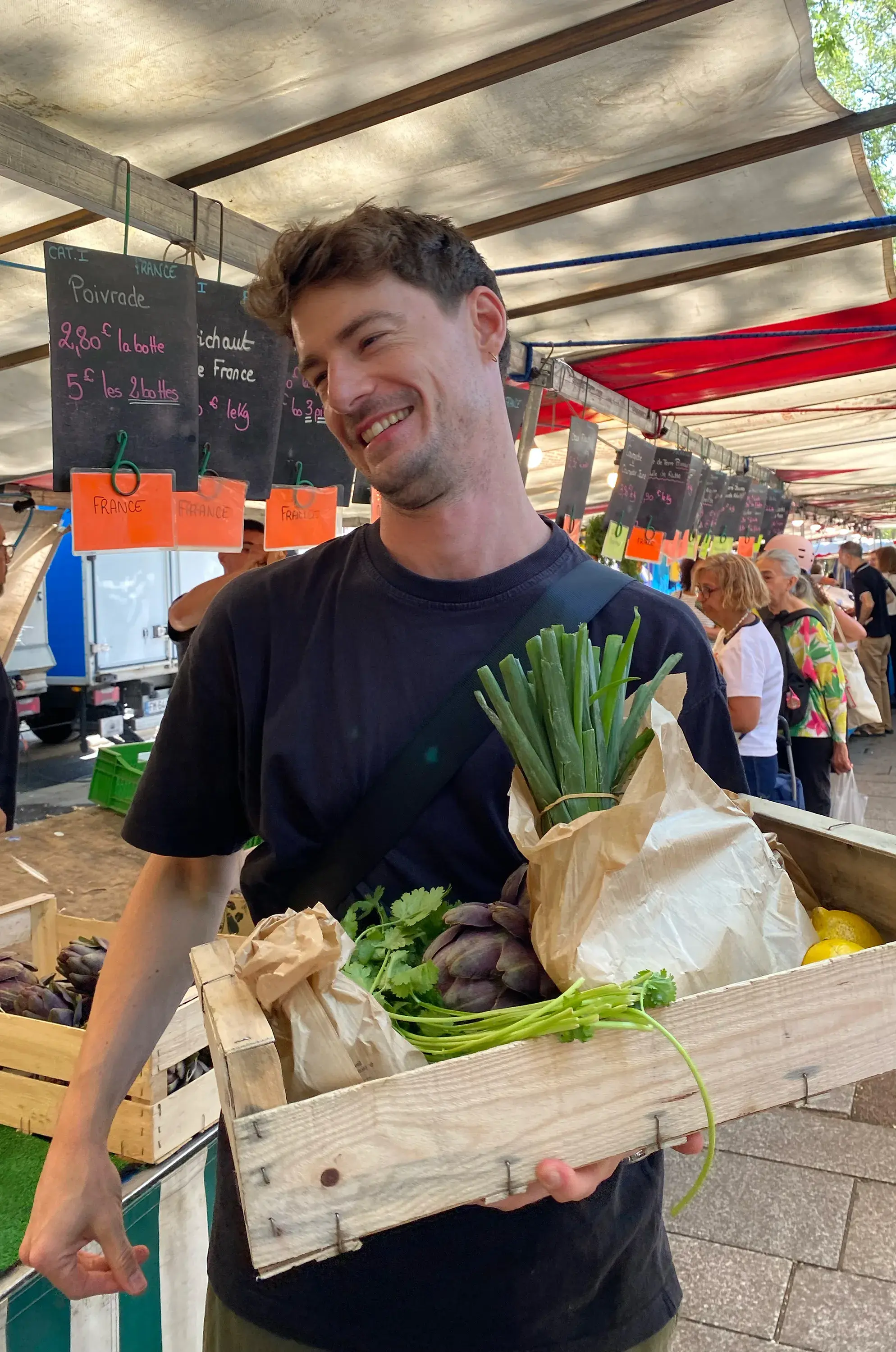 A person smiles while holding a wooden crate filled with fresh vegetables, including green onions and artichokes, at an outdoor market.