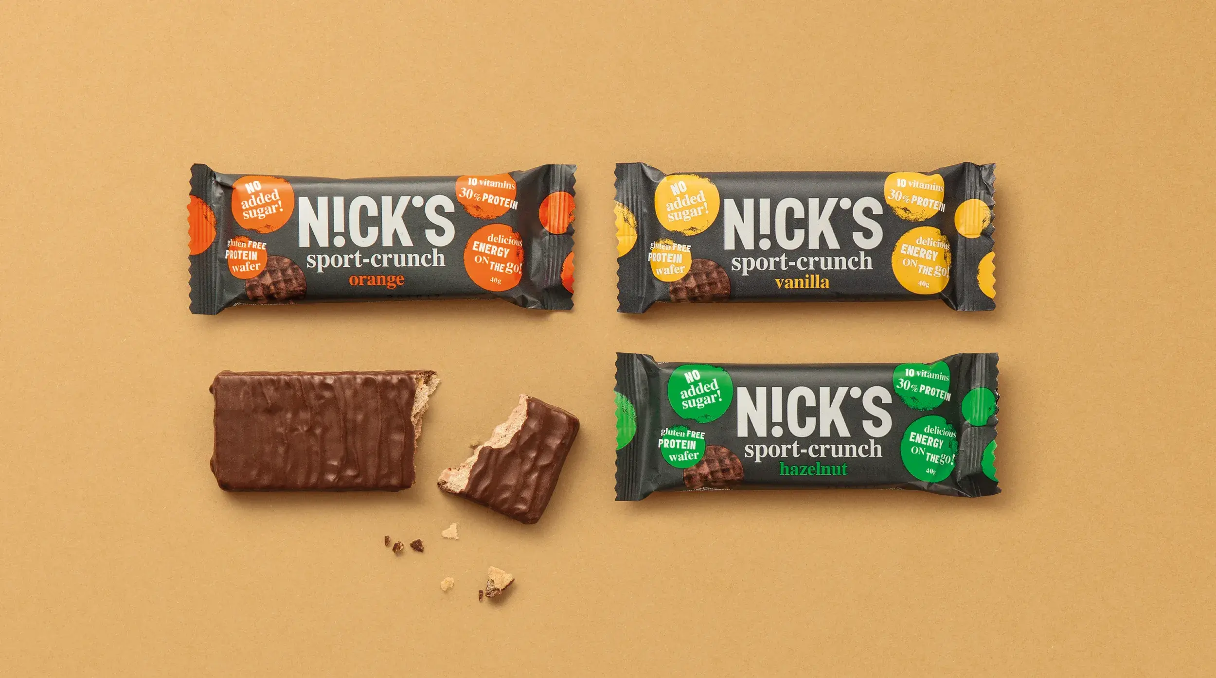 Three Nick's Sport-Crunch bars in orange, vanilla, and hazelnut flavors on a brown surface, with one bar partially unwrapped and bitten.