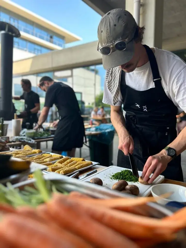 Chef in a black apron and cap chops herbs at an outdoor cooking event, with corn and carrots visible in the foreground.