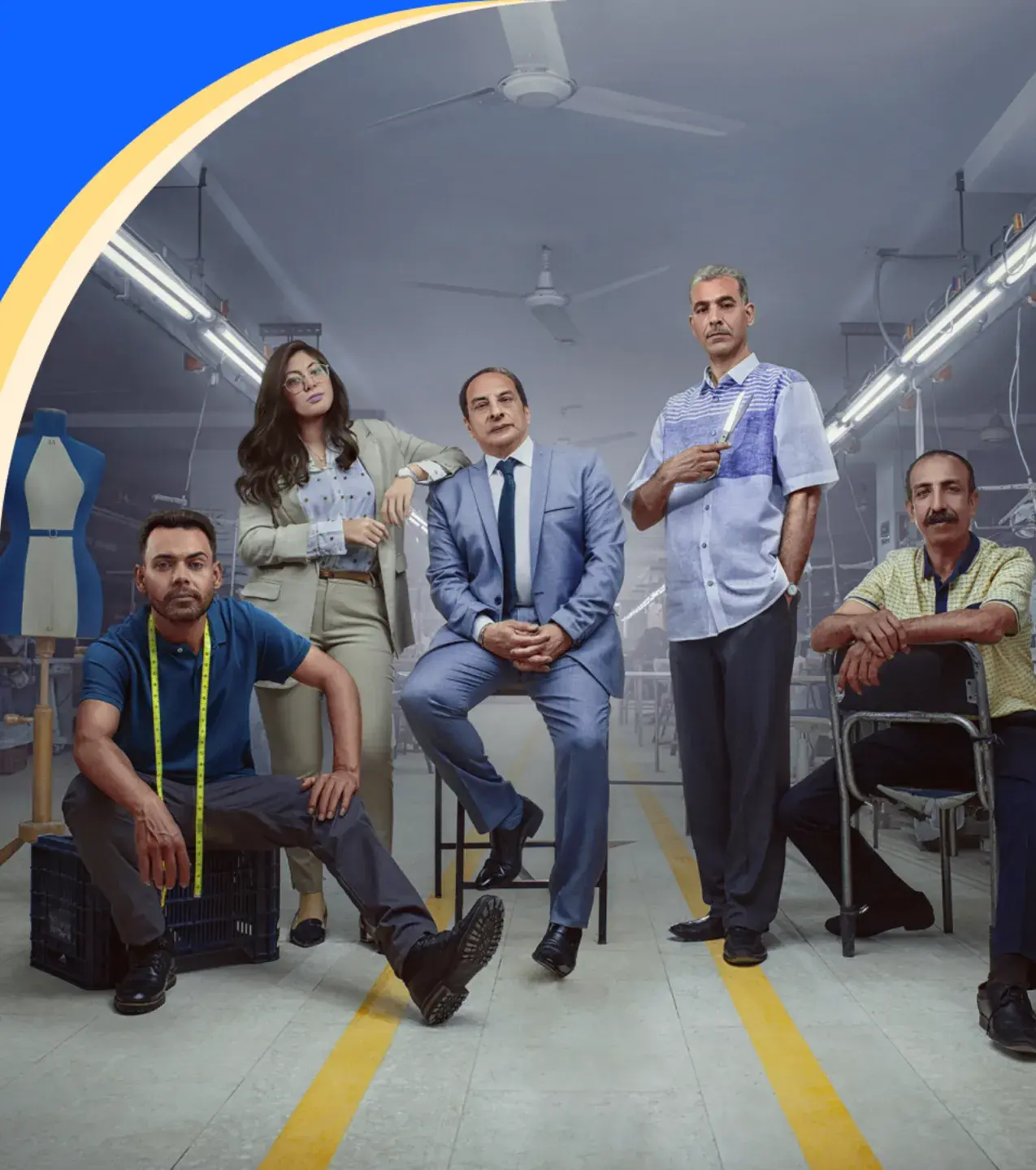 Five people in a tailor shop, with one seated in a suit and others standing or sitting casually, surrounded by sewing equipment.