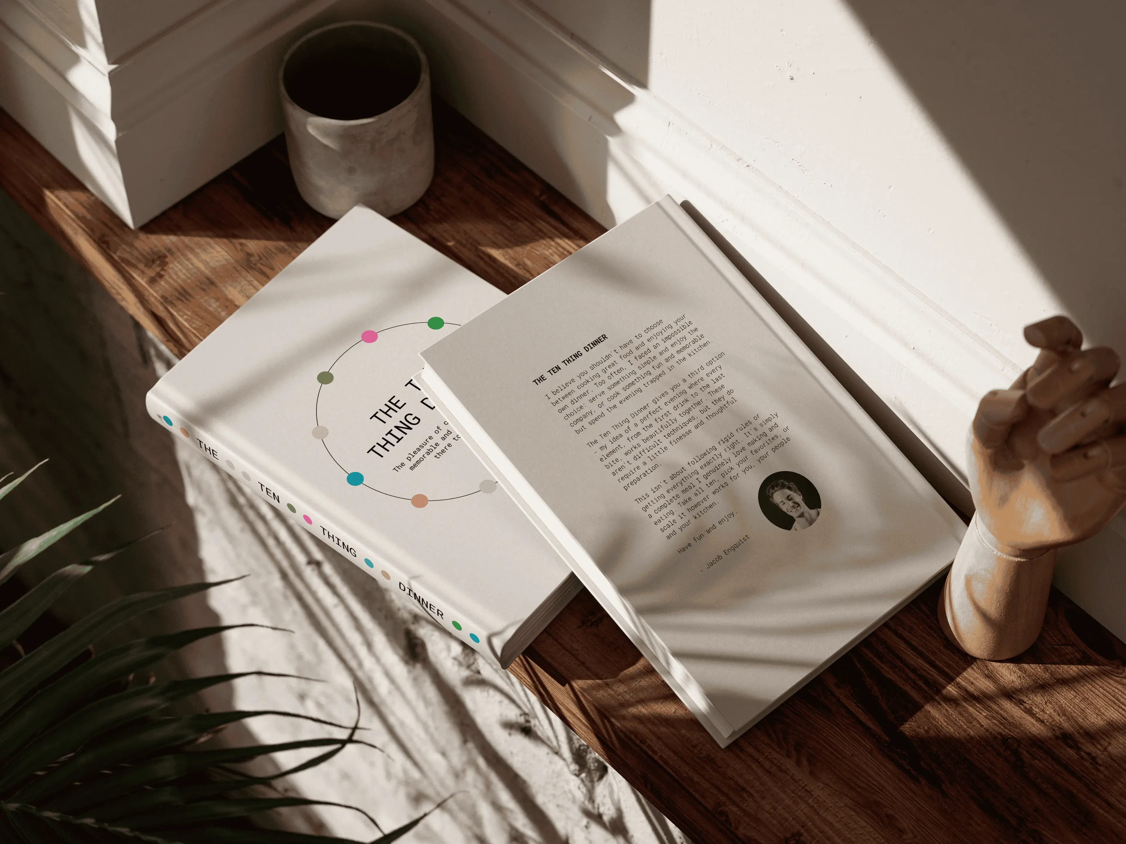 Two books on a wooden surface, one open with text visible. A ceramic cup and a wooden hand sculpture are nearby, with sunlight casting shadows.